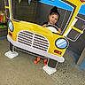 Aurora is registered to the contest to win money with this photo: black_clothing, blue_wall, cardboard_cutout, child, concrete_floor, curious, decor, floor, fun, indoor, mischievous, orange_socks, person, playful, portrait, smile, steering_wheel, toy, wooden_cabinet, yellow_car