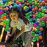 activity, ball_pit, bright_colors, casual_clothing, child, colorful_balls, curly_hair, entertainment, fun, gray_pants, indoor_play_area, leisure, neon_lights, pigtails, play, playground, reflection, slide, white_shirt, young_child