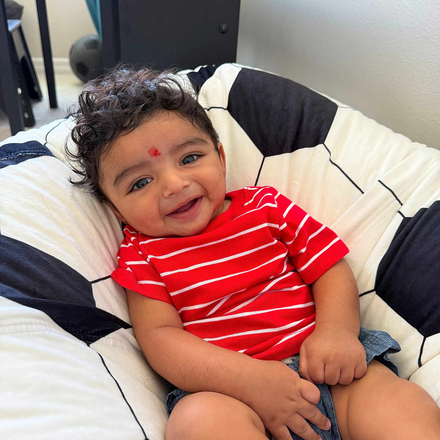 Krishiv joined the competition — help win amazing prizes! baby, child, smiling, curly_hair, red_shirt, striped_clothing, denim_shorts, cushion, soccer_ball_pattern, indoor, furniture, happy, face, person, seated, cute, infant, home, portrait, cozy