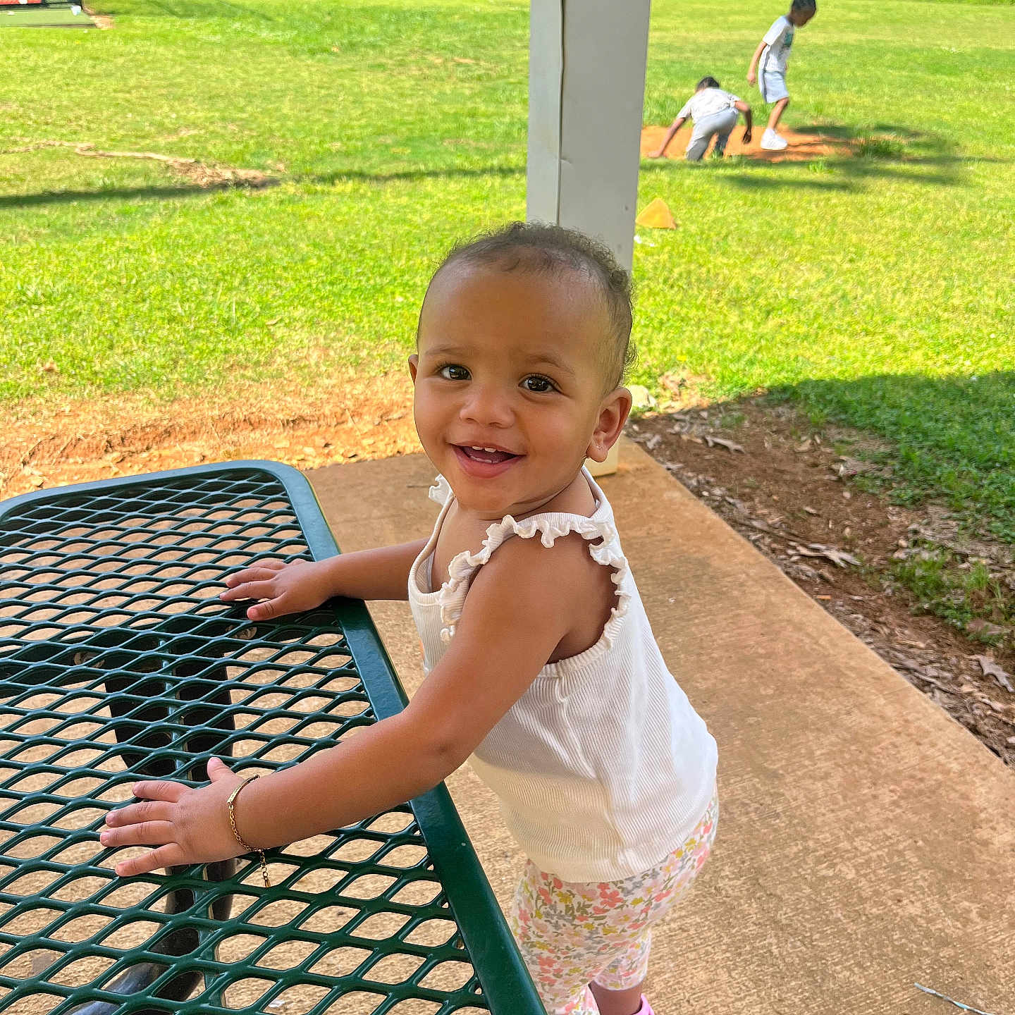 Lailah is registered to the contest to win money with this photo: baseball_field, bracelet, child, concrete, daylight, floral_leggings, grass, greenery, happy, metal_table, outdoor, park, picnic_table, pink_shoes, playing_children, smiling, standing, sunlight, toddler, white_tank_top