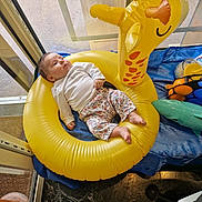 Jade participe au concours pour gagner de l'argent avec cette photo : baby, inflatable, giraffe, yellow, toy, indoor, child, relaxed, blue_cloth, floor, window, natural_light, marble_floor, clothing, patterned_pants, white_shirt, plastic_toys, footwear_absent, face, person