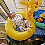 baby, inflatable, giraffe, yellow, toy, indoor, child, relaxed, blue_cloth, floor, window, natural_light, marble_floor, clothing, patterned_pants, white_shirt, plastic_toys, footwear_absent, face, person