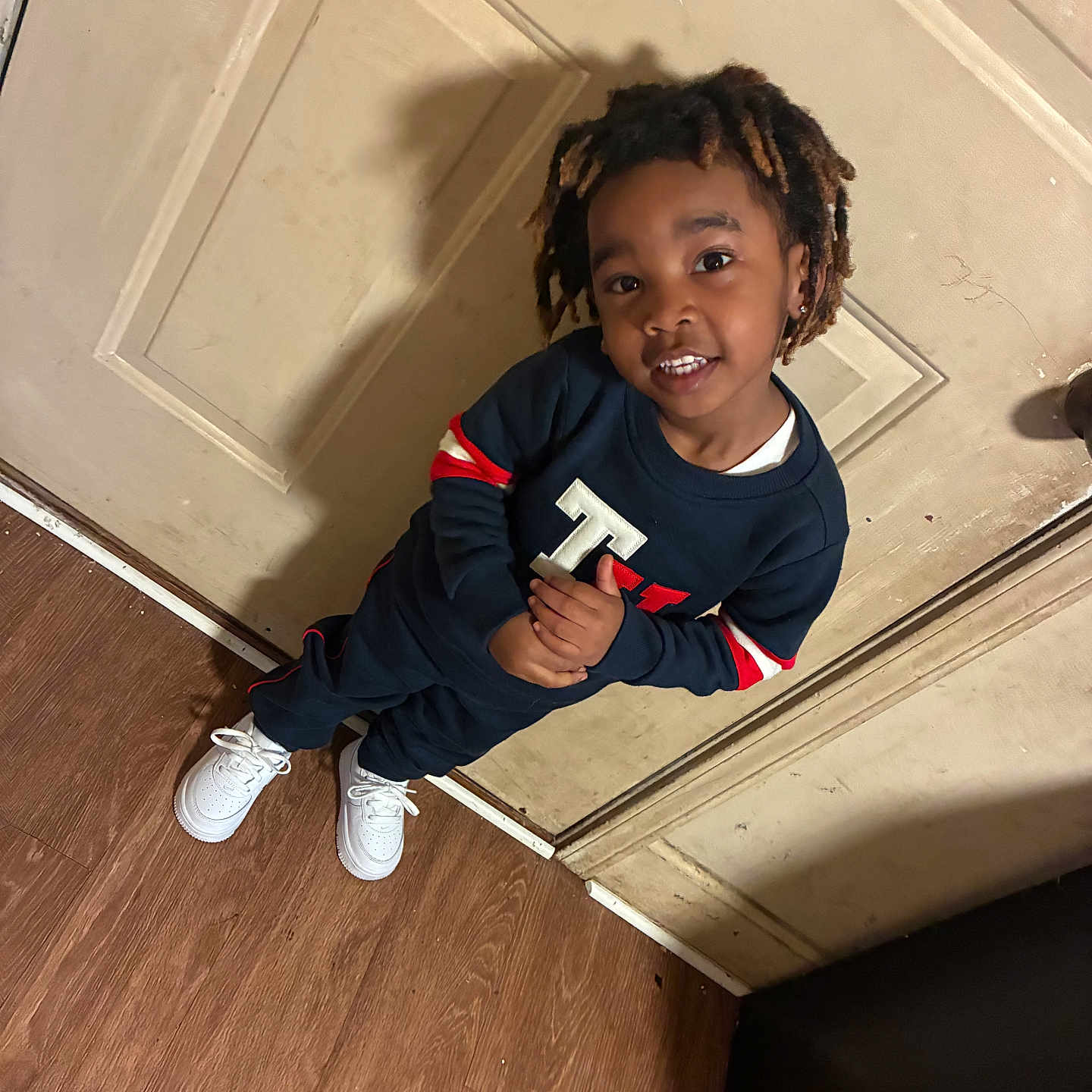 Vamir is registered to the contest to win money with this photo: child, smiling, standing, indoor, door, wooden_floor, casual_clothing, sneakers, hairstyle, person, young, happy, fashion, portrait, playful, confident, home, shadow, floor, wall