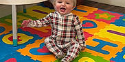 Cruze is registered to the contest to win money with this photo: baby, plaid_pajamas, alphabet_mat, foam_mat, baby_bottle, mickey_mouse, toy, table, clothes, storage_bin, football_shaped, floor, wood_floor, corner, room, happy, smiling, sitting, child, indoor