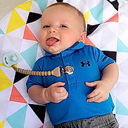 Cruze is registered to the contest to win money with this photo: baby, child, infant, tongue_out, blue_shirt, gray_pants, pacifier, colorful_mat, geometric_pattern, playful, cute, smiling, lying_down, indoors, young_child, person, clothing, hand, face, happy