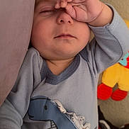 Jacob joined the competition — help win amazing prizes! baby, child, sleepy, onesie, dinosaur, hand, face, infant, couch, plush_toy, blue_clothing, cute, resting, indoors, soft_texture, portrait, young_child, napping, cozy, person