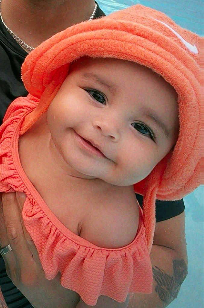 Miyla joined the competition — help win amazing prizes! adult_hand, baby, baby_clothes, child, closeup, cute, face, happy, headwear, holding, infant, orange_clothing, orange_hat, outdoor, person, portrait, skin, smiling, tattoo, water