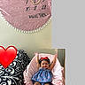 baby, child, smiling, blue_dress, red_bow, pink_chair, pink_blanket, calendar, heart_shape, raised_hands, indoor, cushion, happy, person, furniture, decor, pattern, footwear_free, cute, red_heart_emoji