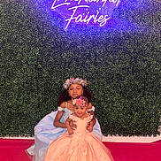 Keymoni is registered to the contest to win money with this photo: child, children, girl, dress, flower_crown, fairy_tale, costume, green_wall, neon_sign, pink_carpet, portrait, smiling, happy, indoor, event, celebration, cute, sitting, fashion, youth
