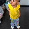 toddler, child, yellow_top, jeans, colorful_pants, indoor, floor, curious, looking_up, shoes, bow_shoes, earrings, standing, clothing, bags, hanging_clothes, door, black_floor, casual, portrait