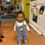 Keymoni is registered to the contest to win money with this photo: adult, chair, child, daycare, denim, flooring, flowers, furniture, happy, indoor, kid, overalls, person, photography, playroom, smiling, toddler, walking, wall_art, yellow_wall