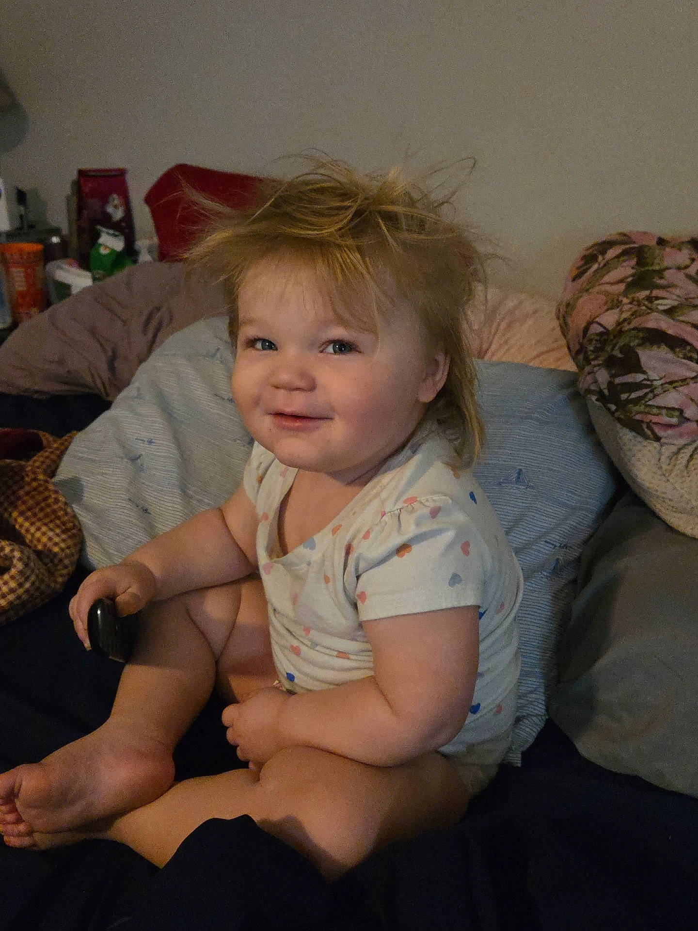 Cheyenne is registered to the contest to win money with this photo: baby, bedroom, blanket, candid, child, chubby, cute, furniture, home, indoor, messy_hair, pajamas, person, pillow, portrait, relaxed, remote_control, sitting, smile, toddler
