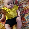 baby, black_shorts, casual_clothing, child, chubby_cheeks, colorful, curious, cute, human, indoors, infant, one_person, patterned_blanket, portrait, relaxed, resting, sitting, soft_lighting, yellow_shirt, young_child