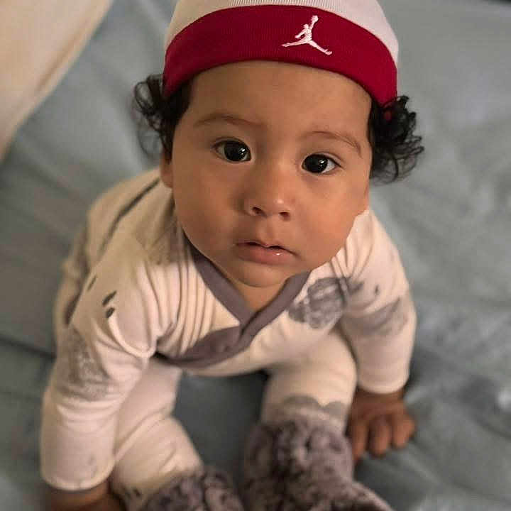 Noah Rios is registered to the contest to win money with this photo: baby, bed, blue_bedspread, boots, cap, child, close_up, curious, cute, fuzzy_boots, hat, indoors, infant, looking_up, pajamas, person, portrait, sitting, soft_lighting, young_child