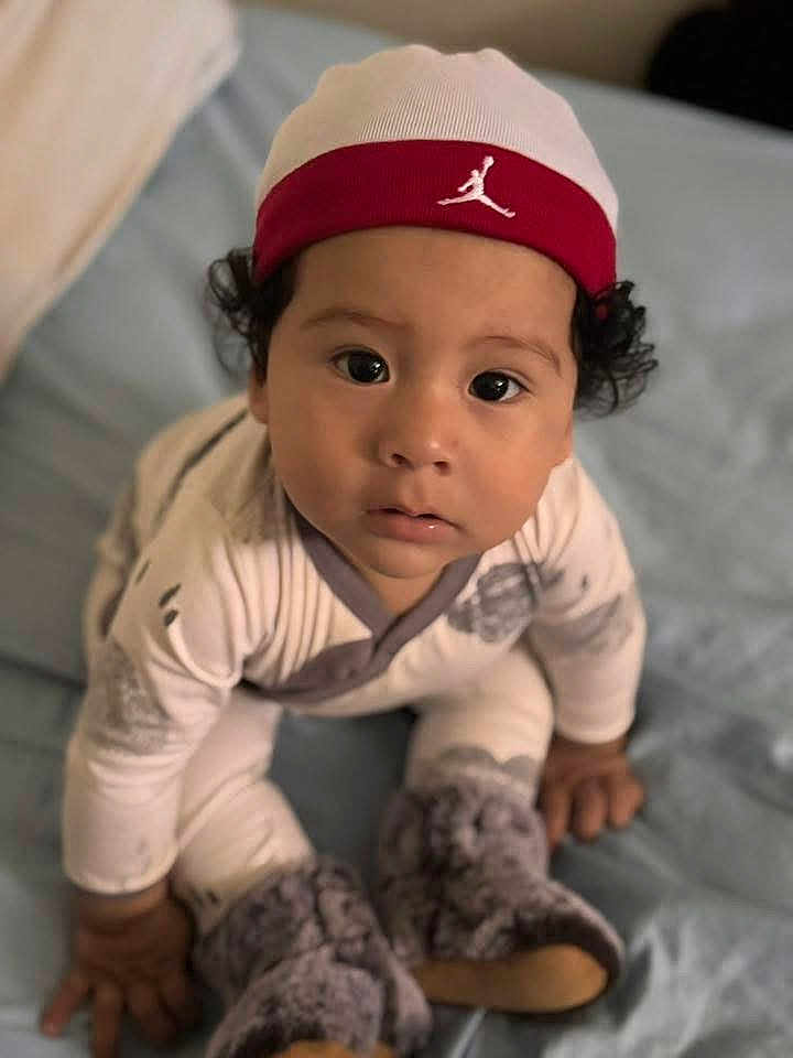 Noah Rios is registered to the contest to win money with this photo: baby, bed, blue_bedspread, boots, cap, child, close_up, curious, cute, fuzzy_boots, hat, indoors, infant, looking_up, pajamas, person, portrait, sitting, soft_lighting, young_child