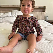 Emma is registered to the contest to win money with this photo: toddler, child, bed, flower_headband, floral_top, denim_shorts, tongue_out, cute, indoor, happy, person, skin, head, face, legs, barefoot, furniture, pillow, blanket, room