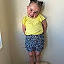 toddler, child, smiling, standing, barefoot, yellow_top, floral_shorts, hair_buns, wooden_stool, indoor, wall, cute, happy, person, portrait, young_child, fashion, playful, skin, head