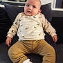 Timaël a rejoint le concours — aidez-le/la à gagner de superbes lots ! baby, infant, child, sitting, couch, pillow, sweater, dinosaur_pattern, brown_pants, white_socks, indoors, cute, chubby_cheeks, portrait, comfortable, cozy, clothing, young_child, curious, smiling