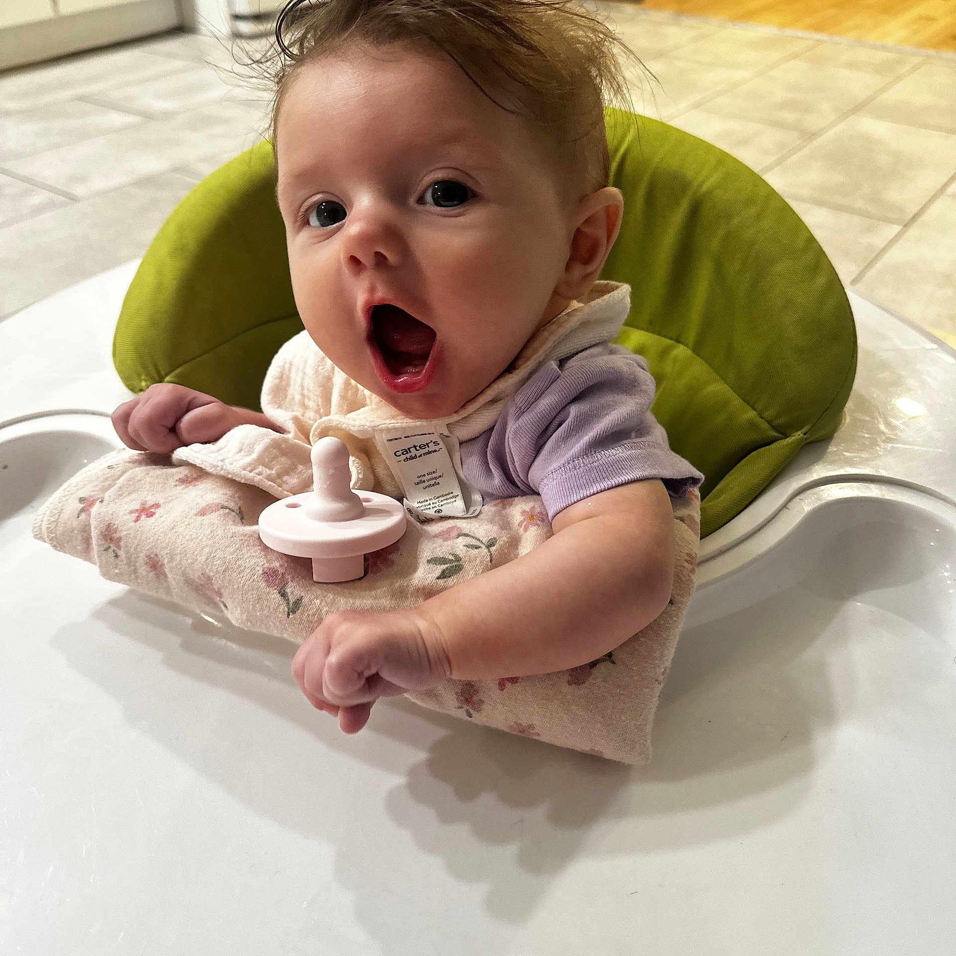 Brielle joined the competition — help win amazing prizes! baby, child, infant, portrait, sitting, booster_seat, highchair, pacifier, bib, floral_clothing, purple_shirt, tousled_hair, wide_eyed, open_mouth, indoor, kitchen, tile_floor, wood_floor, furniture, funny_expression