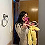 woman, baby, mirror, selfie, bathroom, door, yellow_onesie, smile, phone, long_hair, towel_ring, light_switch, countertop, reflection, clothing, indoor, person, cute, holding, portrait