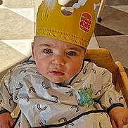 Lévi a rejoint le concours — aidez-le/la à gagner de superbes lots ! baby, child, burger_king, crown, high_chair, bib, indoor, blue_eyes, curious, person, clothing, floor, chair, table, restaurant, pacifier, light, shadow, wood, pattern