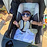 baby, stroller, sunglasses, child, outdoor, sunlight, blanket, toy, person, footwear, sand, hand, clothing, seat, head, smiling, tattoo, accessory, sunshine, relaxation