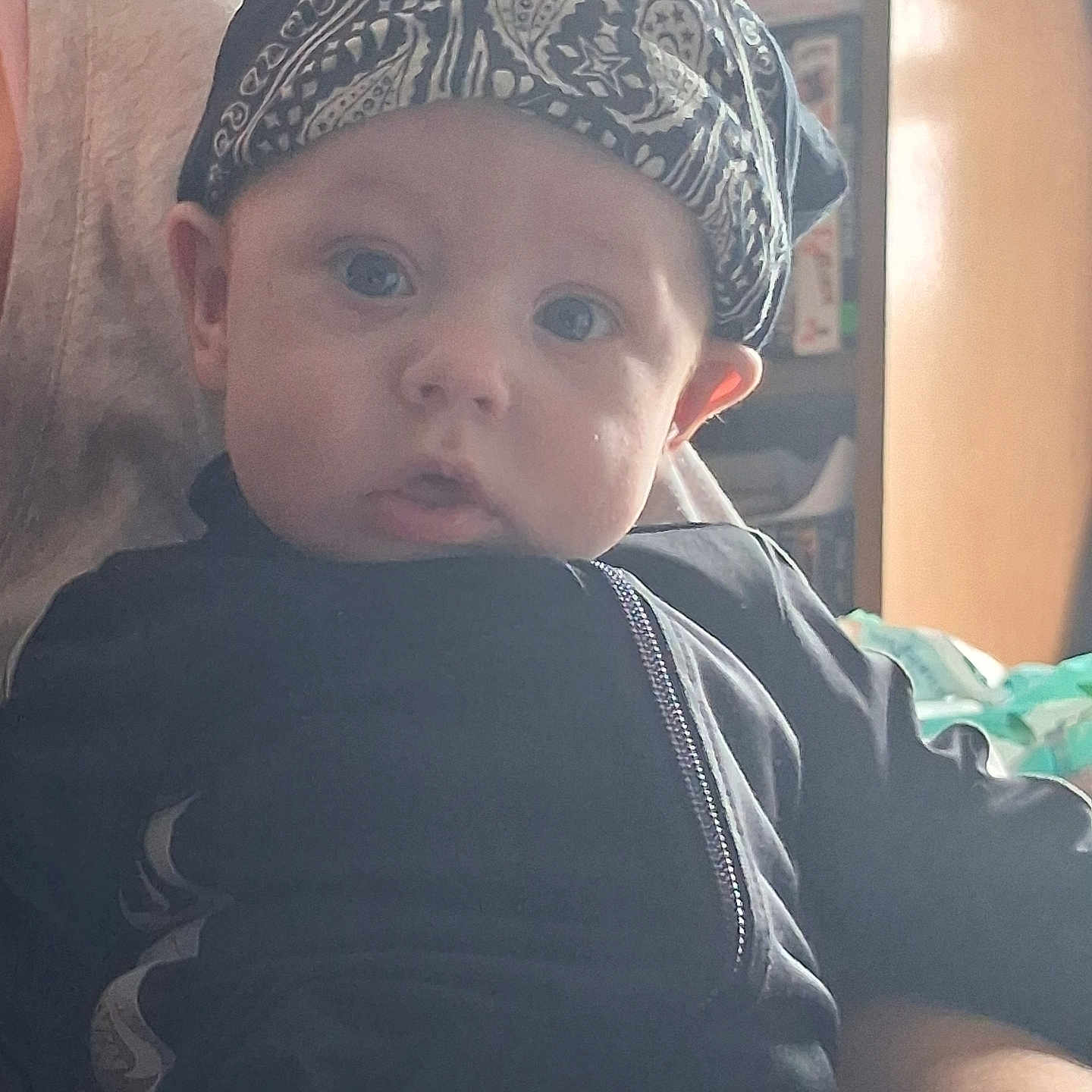 Carter is registered to the contest to win money with this photo: baby, infant, child, portrait, face, eyes, serious_expression, cap, paisley_pattern, black_clothing, jacket, zipper, sitting, indoor, sunlight, chair, home, background, close_up, person