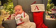 Alexander is registered to the contest to win money with this photo: baby, armchair, christmas_tree, christmas_stocking, holiday, pajamas, bib, ornaments, presents, carpet, lighting, indoor, festive, cozy, smiling, child, red, gray, decorations, celebration
