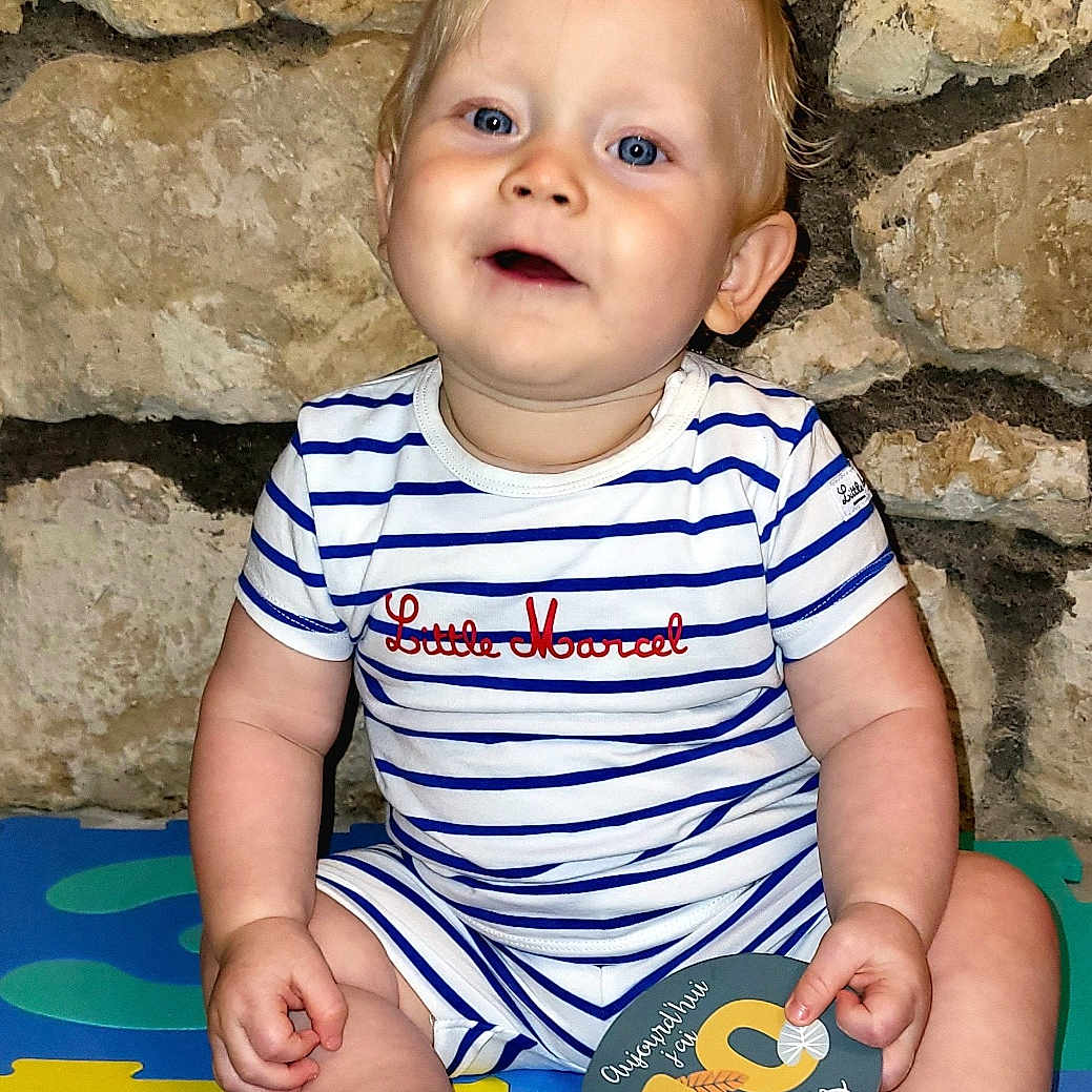 Liam a rejoint le concours — aidez-le/la à gagner de superbes lots ! baby, barefoot, blue_eyes, child, chubby_cheeks, colorful_mats, cute, foam_puzzle, happy, indoor, infant, milestone_card, onesie, person, playtime, portrait, sitting, smiling, stone_wall, striped_clothing