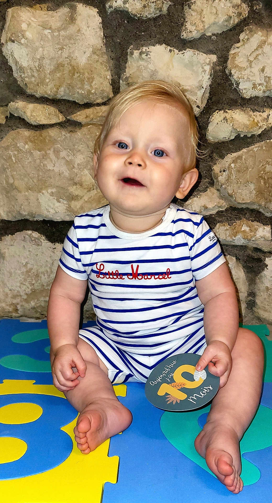 Liam a rejoint le concours — aidez-le/la à gagner de superbes lots ! baby, child, blue_eyes, striped_clothing, onesie, milestone_card, sitting, barefoot, colorful_mats, foam_puzzle, stone_wall, indoor, happy, cute, smiling, portrait, infant, chubby_cheeks, playtime, person