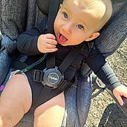 Brooklyn is registered to the contest to win money with this photo: baby, child, stroller, seatbelt, outdoor, sunlight, blue_eyes, blond_hair, black_clothing, cute, infant, hand, smiling, happy, person, legs, face, baby_boy, daylight, casual