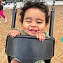 toddler, child, swing, playground, smiling, curly_hair, happy, outdoor, park, wood_chips, bench, person, baby_swing, hand, face, clothing, green_sweater, shorts, daylight, fun