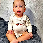 Charly participe au concours pour gagner de l'argent avec cette photo : baby, blanket, blue_eyes, child, clothing, curious, cute, expression, feet, fingers, indoor, infant, onesie, person, portrait, sitting, skin, soft_texture, toddler, wall