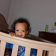 Octavius is registered to the contest to win money with this photo: toddler, crib, child, smile, blue_shirt, hand, furniture, bedroom, dresser, indoor, curly_hair, person, baby, face, young_child, wooden_crib, room, home, cute, standing