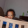 toddler, crib, child, smile, blue_shirt, hand, furniture, bedroom, dresser, indoor, curly_hair, person, baby, face, young_child, wooden_crib, room, home, cute, standing