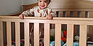 Octavius is registered to the contest to win money with this photo: child, toddler, baby, crib, wooden_crib, blanket, plaid_blanket, pajamas, football_print, standing, smiling, face, stuffed_toy, plush, mattress, nursery, indoor, bars, cute, portrait