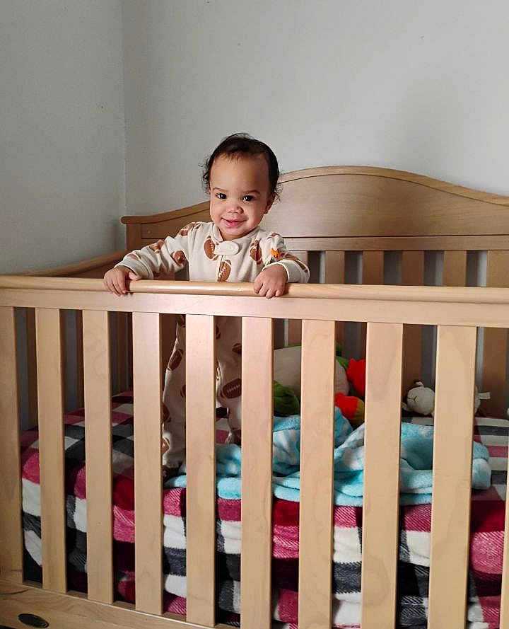 Octavius is registered to the contest to win money with this photo: child, toddler, baby, crib, wooden_crib, blanket, plaid_blanket, pajamas, football_print, standing, smiling, face, stuffed_toy, plush, mattress, nursery, indoor, bars, cute, portrait