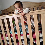 Octavius is registered to the contest to win money with this photo: baby, infant, crib, wooden_crib, blanket, plaid_blanket, blue_blanket, pajamas, baby_bed, toddler, child, face, smile, hand, railing, toy, pacifier_clip, furniture, interior, portrait