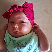 Lily-Rose is registered to the contest to win money with this photo: baby, infant, headband, pink_bow, green_shirt, cartoon_print, face, cheeks, hand, curious, lying_down, closeup, skin, eyes, expression, indoors, texture, fabric, person, child