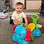 toddler, child, toy, elephant_toy, indoor, carpet, cabinet, book, play, smile, person, furniture, floor, plastic_toy, colorful, seat, wooden, happy, room, baby