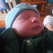 Jordan is registered to the contest to win money with this photo: baby, sleeping, hat, green_clothing, indoor, infant, face, peaceful, cute, soft_light, wooden_furniture, drawer, blurred_background, hand, slippers, cozy, resting, child, portrait, closeup