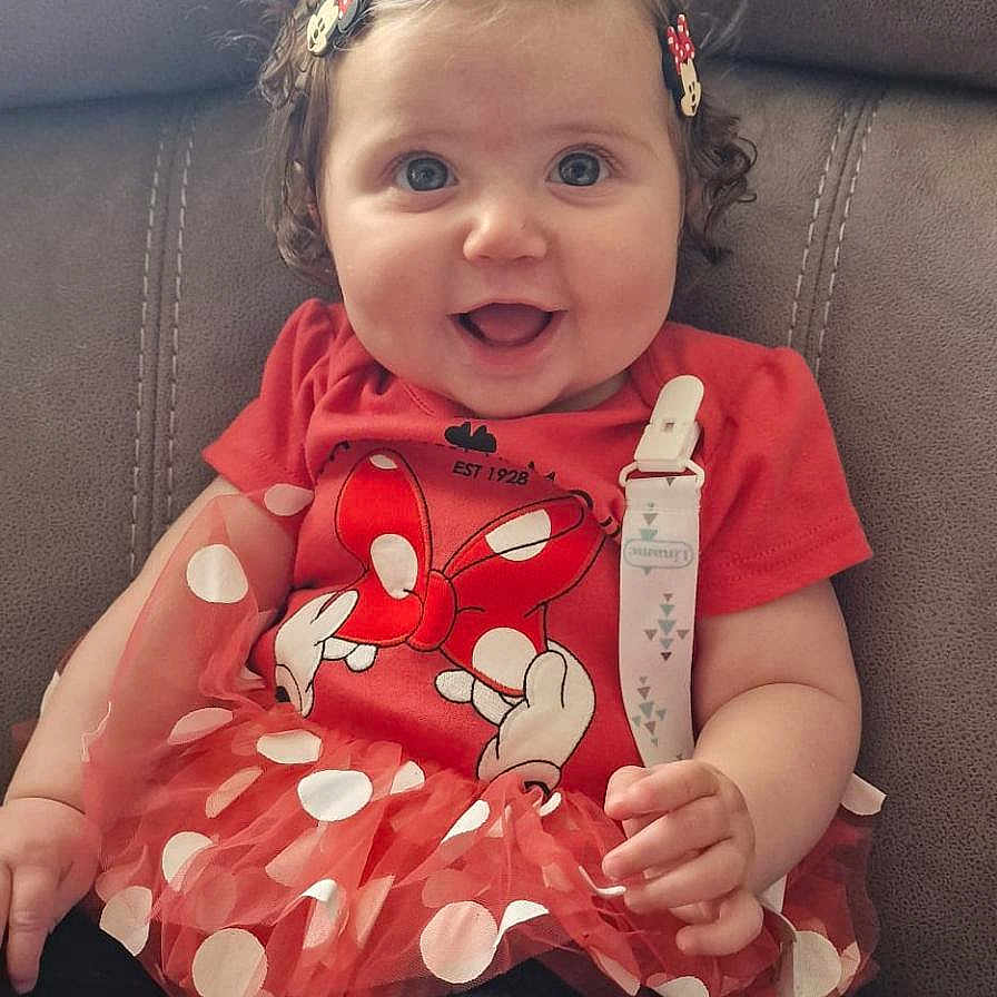 Esme joined the competition — help win amazing prizes! baby, black_leggings, child, clothing, couch, curly_hair, cute, face, fun, hair_clips, happy, indoor, minnie_mouse, person, polka_dot, portrait, red_dress, sitting, smiling, toddler