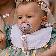 Giulia participe au concours pour gagner de l'argent avec cette photo : baby, pacifier, headband, flower_crown, bib, dress, daisy_pattern, child, infant, person, closeup, face, hand, clothing, accessory, necklace, adult, skin, cute, expression