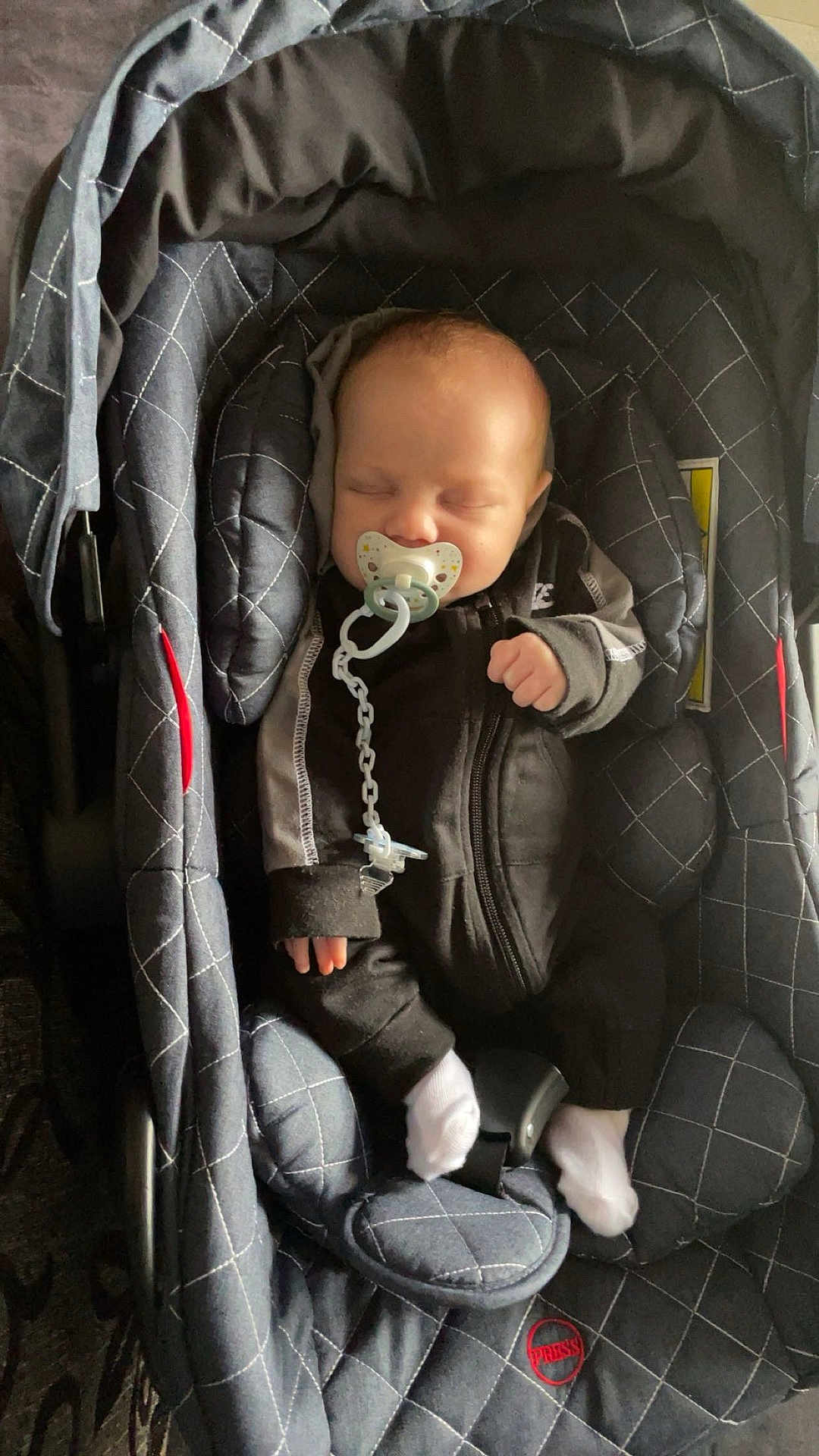 Marlon a rejoint le concours — aidez-le/la à gagner de superbes lots ! baby, infant, sleeping_baby, pacifier, pacifier_chain, car_seat, infant_carrier, buckle, sock, zipper, hooded_clothing, hand, head, quilted_pattern, dark_clothing, cozy, portrait, newborn, indoor, soft_lighting