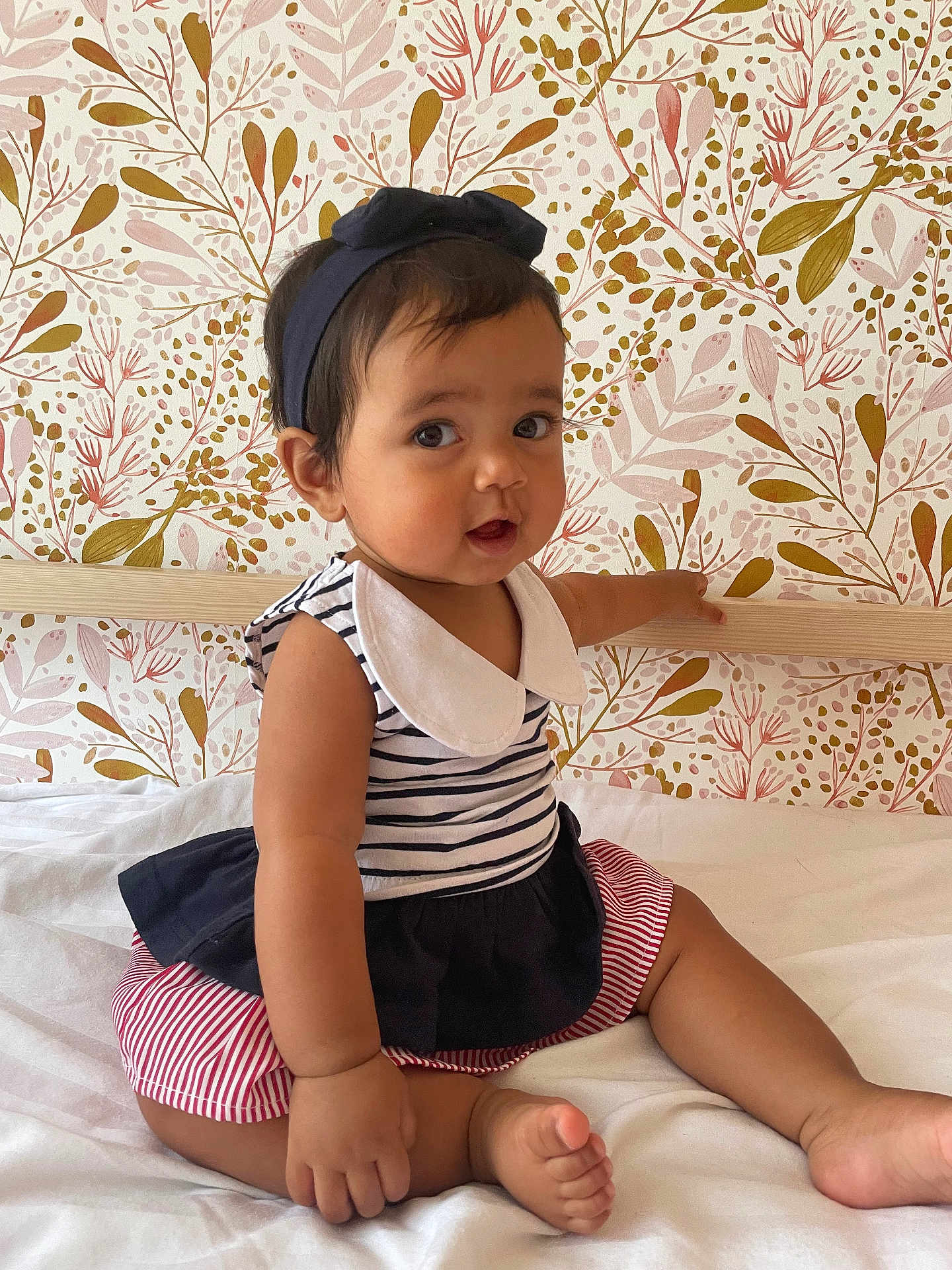 Calypso participe au concours pour gagner de l'argent avec cette photo : baby, child, infant, sitting, bed, striped_clothing, headband, curly_hair, cute, floral_wallpaper, white_bed, indoors, portrait, skin, barefoot, pink, navy_blue, red_stripes, collar, expression
