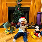 Kian joined the competition — help win amazing prizes! child, toddler, santa_hat, christmas_tree, fireplace, stuffed_frog, mickey_mouse, plush_toy, hardwood_floor, wrapped_gifts, gold_ribbon, ornaments, mantel, holiday, smiling, indoor, sitting, toy, cozy, decorations