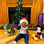 child, toddler, santa_hat, christmas_tree, fireplace, stuffed_frog, mickey_mouse, plush_toy, hardwood_floor, wrapped_gifts, gold_ribbon, ornaments, mantel, holiday, smiling, indoor, sitting, toy, cozy, decorations