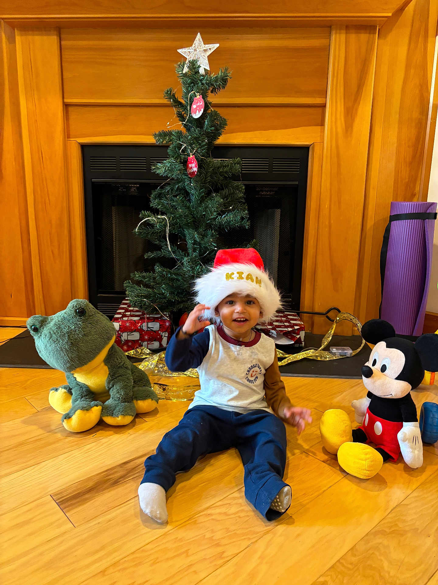 Kian joined the competition — help win amazing prizes! child, toddler, santa_hat, christmas_tree, fireplace, stuffed_frog, mickey_mouse, plush_toy, hardwood_floor, wrapped_gifts, gold_ribbon, ornaments, mantel, holiday, smiling, indoor, sitting, toy, cozy, decorations