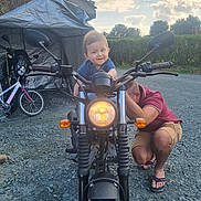 Diego participe au concours pour gagner de l'argent avec cette photo : toddler, motorcycle, adult, smiling, outdoor, gravel, bicycle, daytime, cloudy_sky, vehicle, person, casual_clothing, headlight, nature, hedge, footwear, crouching, happy, transport, garage