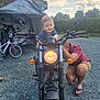 toddler, motorcycle, adult, smiling, outdoor, gravel, bicycle, daytime, cloudy_sky, vehicle, person, casual_clothing, headlight, nature, hedge, footwear, crouching, happy, transport, garage